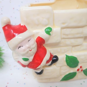 May include: A ceramic Christmas planter in the shape of a chimney, with a Santa Claus figure climbing the side. The planter is cream-colored with brick detailing, and Santa is wearing a red suit and hat with green accents.