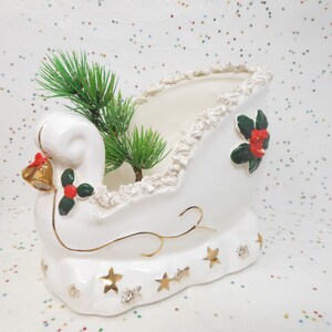 May include: A white ceramic sleigh-shaped planter with gold accents and a ruffled rim. The sleigh features gold stars, holly berries, and a bell. Green faux pine branches are inside the planter, creating a festive holiday decoration.