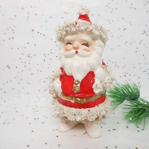 May include: A ceramic Santa Claus figurine with a red coat, white beard, and a matching hat. The figurine has a gold belt and is decorated with cream-colored trim. The background is white with colorful confetti.