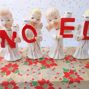 May include: Vintage ceramic Christmas angel figurines holding the letters spelling "NOEL" in red. The angels are dressed in white robes with gold accents and are standing on a box with a poinsettia pattern.