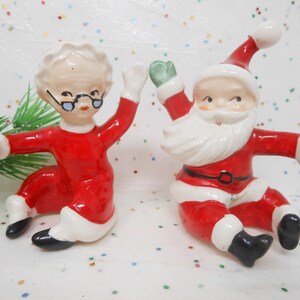 May include: A pair of ceramic Christmas figurines, Santa Claus and Mrs. Claus, in a playful pose. They are dressed in red suits with white trim, black boots, and green gloves. Mrs. Claus wears glasses.
