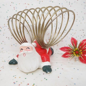 May include: A vintage Christmas card holder featuring a Santa Claus figurine in red, white, and green. The card holder has a gold-colored wire spiral design. A red poinsettia flower is to the right of the card holder.