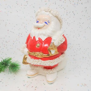 May include: A ceramic Santa Claus figurine. The figure is dressed in a red coat with white trim and a gold belt. Santa has a white beard and a smiling face with blue eyes. He is holding a gold sack.