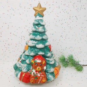 May include: A ceramic Christmas tree with a gold star on top. The green tree is decorated with white accents, red and orange gift boxes, and a teddy bear wearing a Santa hat. A festive holiday decoration.