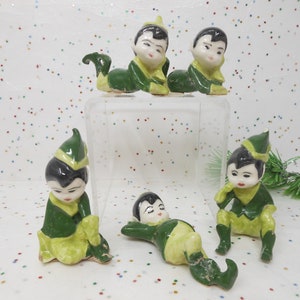 May include: Set of six green and white ceramic elf figurines. The elves are all wearing green hats and have black hair. They are in various poses, including sitting, lying down, and standing.