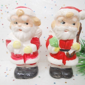 May include: Two vintage ceramic Santa Claus figurines. Each Santa is dressed in a red suit with white trim, a white beard, and a red hat. One holds a bag, the other a staff. Black boots complete the look. A festive holiday decoration.
