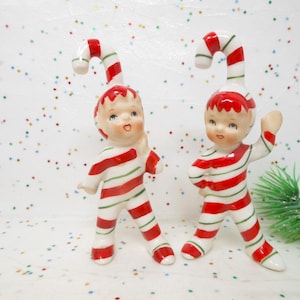 May include: Two ceramic figurines of children in red and white striped outfits, each with a candy cane-shaped hat. The figurines have rosy cheeks and are posed in a playful stance, evoking a festive holiday theme.