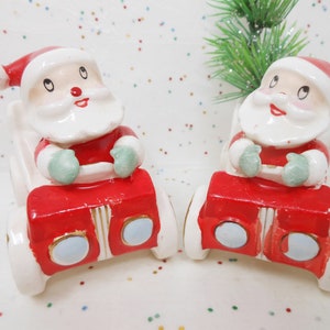 May include: Two ceramic Santa Claus figurines in red suits and hats, seated in red and white sleighs. The Santas have white beards and rosy cheeks, with green mittens. A festive Christmas decoration.