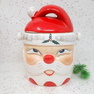 May include: A vintage Santa Claus cookie jar. The jar features a red hat, a white face with rosy cheeks, a red nose, and a smiling mouth. The jar has a white base with red polka dots and gold accents.