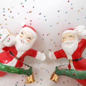 May include: Two ceramic Santa Claus figurines in red suits and white beards, each with a green garland and gold bell. One carries a sack, the other a candy cane. Festive Christmas decor.