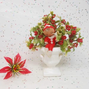 May include: A vintage Christmas decoration featuring a small figurine in a red suit and hat, seated in a white urn filled with holly and red berries. A red and yellow flower is on the left.