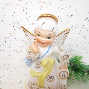 May include: A vintage ceramic angel figurine with white wings, a gold halo, and a yellow ribbon. The angel is holding a small clock and wearing a white dress with gold accents. The figurine is a Christmas or holiday decoration.