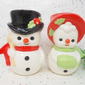 May include: A pair of ceramic snowman figurines. One snowman wears a black top hat and red scarf, holding a yellow shovel. The other snowman has a red hat and green scarf. Both have black eyes, orange noses, and black button details.