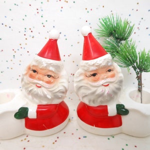 May include: Two ceramic Santa Claus figurines with red hats and coats, white beards, and green gloves. Each holds a white sack-shaped container, with a small green pine branch in one. Festive Christmas decor.