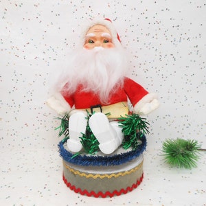 May include: A vintage Santa Claus figurine with a white beard, red suit, and white boots. The Santa figure sits atop a round, decorated base with blue and gold trim, and green garland. A festive Christmas decoration.