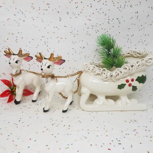 May include: A vintage Christmas decoration featuring two white ceramic reindeer pulling a sleigh. The reindeer have gold antlers and are connected by a gold chain. The sleigh is decorated with holly and a sprig of evergreen.