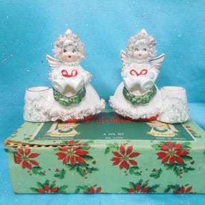May include: Two vintage ceramic angel candle holders. Each angel is white with a ruffled skirt, holding a heart and wearing a green wreath. The candle holders sit on a green box with red poinsettias.