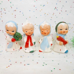 May include: Four vintage ceramic angel figurines. Each angel wears a white robe with red and green accents, and a colored scarf. One angel holds a green wreath, another a red bird, and the others have their hands clasped.