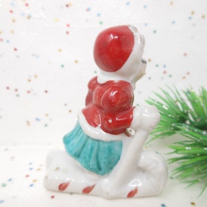 K&ouml;nnte beinhalten: Kleine Keramikfigur einer Person in rotem Mantel und Hut, die einen wei&szlig;en Schlitten f&auml;hrt. Der Schlitten hat rot-wei&szlig;e Zuckerstangenstreifen. Die Person tr&auml;gt einen t&uuml;rkisfarbenen Rock. Ein Zweig gr&uuml;ner, glitzernder Kiefer im Hintergrund.