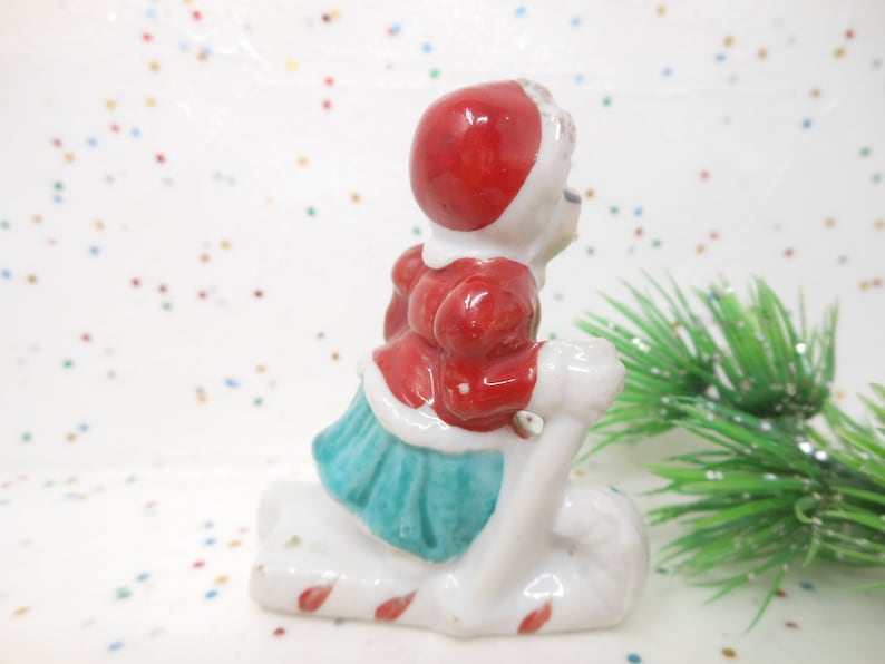 K&ouml;nnte beinhalten: Kleine Keramikfigur einer Person in rotem Mantel und Hut, die einen wei&szlig;en Schlitten f&auml;hrt. Der Schlitten hat rot-wei&szlig;e Zuckerstangenstreifen. Die Person tr&auml;gt einen t&uuml;rkisfarbenen Rock. Ein Zweig gr&uuml;ner, glitzernder Kiefer im Hintergrund.