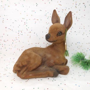 May include: A brown, velvet-textured deer figurine in a resting pose. The deer has large ears, a black nose, and detailed eyes. A small sprig of green foliage is attached to its neck. The figurine is a decorative item.