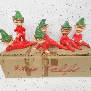 May include: A collection of vintage ceramic elf figurines in red suits and green hats. The elves are posed in various positions on top of a cardboard box. The box has the word "Xmas" and "Elfes" written on it in red.