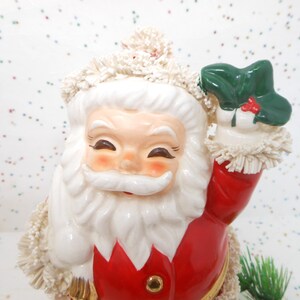 May include: A ceramic Santa Claus figurine with a smiling face, white beard, and red suit. The figurine is holding a green holly sprig with red berries. The Santa Claus has a white hat and trim.
