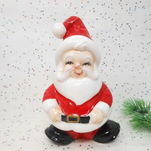 May include: A ceramic Santa Claus figurine, featuring a red suit, white beard, and a matching red hat with a white pom-pom. The figurine has black boots and a black belt with a gold buckle. The Santa is smiling.