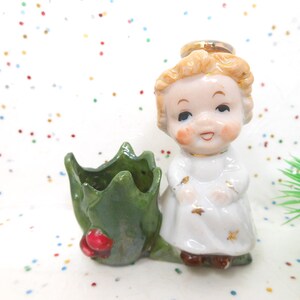 May include: A vintage ceramic angel figurine with a gold halo, wearing a white dress with gold accents. The angel is next to a green leaf-shaped holder with a red berry. A charming Christmas decoration.