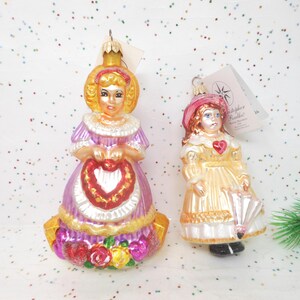 May include: Two glass Christmas ornaments. One is a woman in a purple dress holding a heart. The other is a girl in a yellow dress holding an umbrella. Both have gold accents and are vintage.
