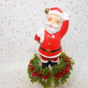 May include: A ceramic Santa Claus figurine in a red suit with a white beard and hat, waving with one arm raised. The figurine is set on a green holly wreath with red berries, a classic Christmas decoration.