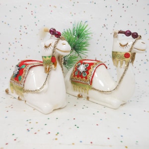 May include: Two white ceramic camel figurines with decorative saddles in red, green, and gold. Each camel has a gold bridle and a decorative headpiece with red beads. The figurines are likely for home decor or a holiday display.