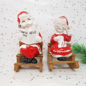 May include: A pair of vintage Christmas figurines. One is Santa Claus reading a newspaper with "North News Pole" printed on it. The other is Mrs. Claus holding a stocking. Both are seated on wooden sleds.