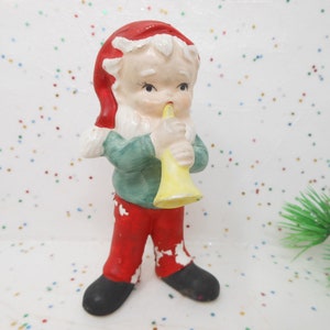 May include: A vintage Christmas figurine of a small elf playing a yellow horn. The elf wears a red hat and pants, a green shirt, and black shoes. The figurine has a painted, aged appearance, suggesting it is an antique holiday decoration.