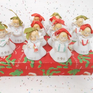 May include: A collection of vintage ceramic angel figurines with white robes, gold wings, and colorful ribbon accents. The angels are arranged on a red box with a Christmas-themed design, perfect for holiday decor.