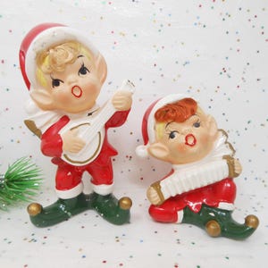 May include: Two vintage ceramic Christmas elf figurines. One elf plays a banjo, the other an accordion. Both wear red and white Santa hats, red suits, green shoes with gold accents, and have blonde hair. The figurines are on a white surface with colorful confetti.