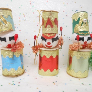 May include: Three vintage Christmas ornaments shaped like toy soldiers. Each ornament features a drum-shaped base with gold trim, a white face with black eyes and a red nose, and a tall hat with gold accents. The ornaments are in blue, red, and green.