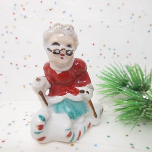 K&ouml;nnte beinhalten: Kleine Keramikfigur einer Frau in rotem und wei&szlig;em Outfit, die eine Brille tr&auml;gt. Sie sitzt auf einem wei&szlig;en Sack mit roten und gr&uuml;nen Streifen und h&auml;lt einen Stock. Ein weihnachtliches Dekorationsst&uuml;ck.