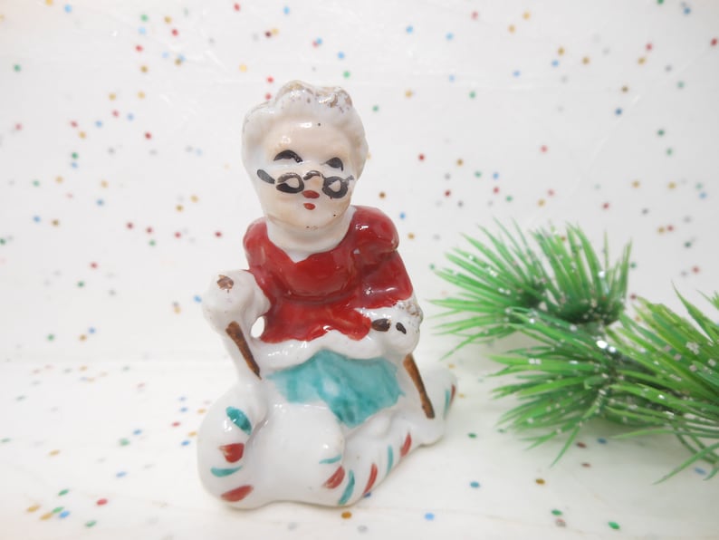 K&ouml;nnte beinhalten: Kleine Keramikfigur einer Frau in rotem und wei&szlig;em Outfit, die eine Brille tr&auml;gt. Sie sitzt auf einem wei&szlig;en Sack mit roten und gr&uuml;nen Streifen und h&auml;lt einen Stock. Ein weihnachtliches Dekorationsst&uuml;ck.