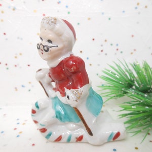 K&ouml;nnte beinhalten: Eine Vintage-Keramikfigur von Mrs. Claus, die einen roten Mantel, einen wei&szlig;en Hut und einen blauen Rock tr&auml;gt. Sie h&auml;lt einen Stock und einen Sack mit einer Zuckerstangenbasis. Die Figur hat goldene Akzente und ist eine Weihnachtsdekoration.