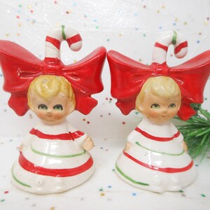 May include: Pair of vintage Christmas figurines. Each figurine is a small girl with blonde hair, wearing a white dress with red and green stripes. A large red bow and candy cane top each figurine. Festive holiday decor.