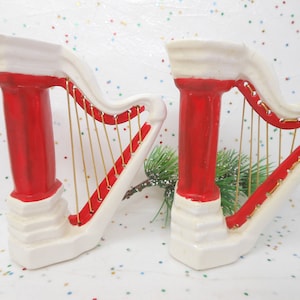 May include: A pair of decorative harp-shaped figurines featuring a glossy white base and pillar with a vibrant red accent on the column and frame. Each harp is strung with thin gold-colored wire, creating a musical instrument aesthetic.