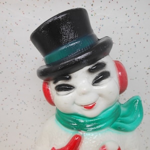 May include: A vintage snowman figurine with a black top hat, green scarf, and red earmuffs. The snowman has a smiling face with black eyes and a red mouth. The figurine is white with a glossy finish.