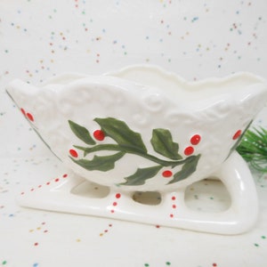 May include: A white ceramic sleigh-shaped dish with a scalloped rim. The dish is decorated with green holly leaves and red berries. The sleigh has runners and is suitable for holiday decor.