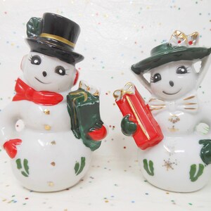 May include: Pair of ceramic snowman figurines. One snowman wears a black top hat and red scarf, holding a green gift. The other snowman wears a green hat and holds a red gift. Both have gold accents and are smiling.