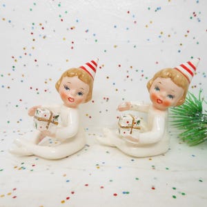 May include: Two ceramic figurines of seated elves, each holding a wrapped gift. The elves have blonde hair, rosy cheeks, and wear white outfits and red and white striped party hats. The background is white with colorful confetti.
