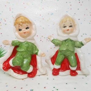 May include: Two ceramic figurines of children in green outfits, white hooded cloaks, and red capes. The figurines have blonde hair and are posed with their arms outstretched. The figures are likely Christmas decorations.