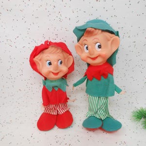 May include: Two vintage elf dolls. One doll wears a red outfit with a matching hood and striped pants. The other doll wears a green outfit with a matching hat and striped pants. Both dolls have rosy cheeks and smiling faces.