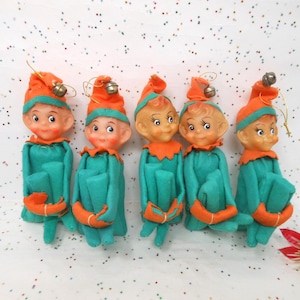 May include: Five vintage Christmas elf ornaments. Each elf has a painted face, orange hat with a bell, and teal felt clothing with orange accents. The ornaments are arranged on a white surface with colorful confetti.