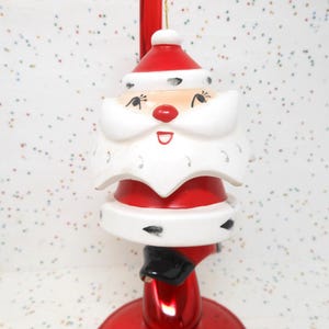 May include: A vintage ceramic Christmas ornament featuring a Santa Claus figure with a white beard and red hat. The ornament is designed with a layered look, with the Santa's face, beard, and hat appearing to be separate pieces. The ornament is attached to a red and white candy cane-shaped stand.